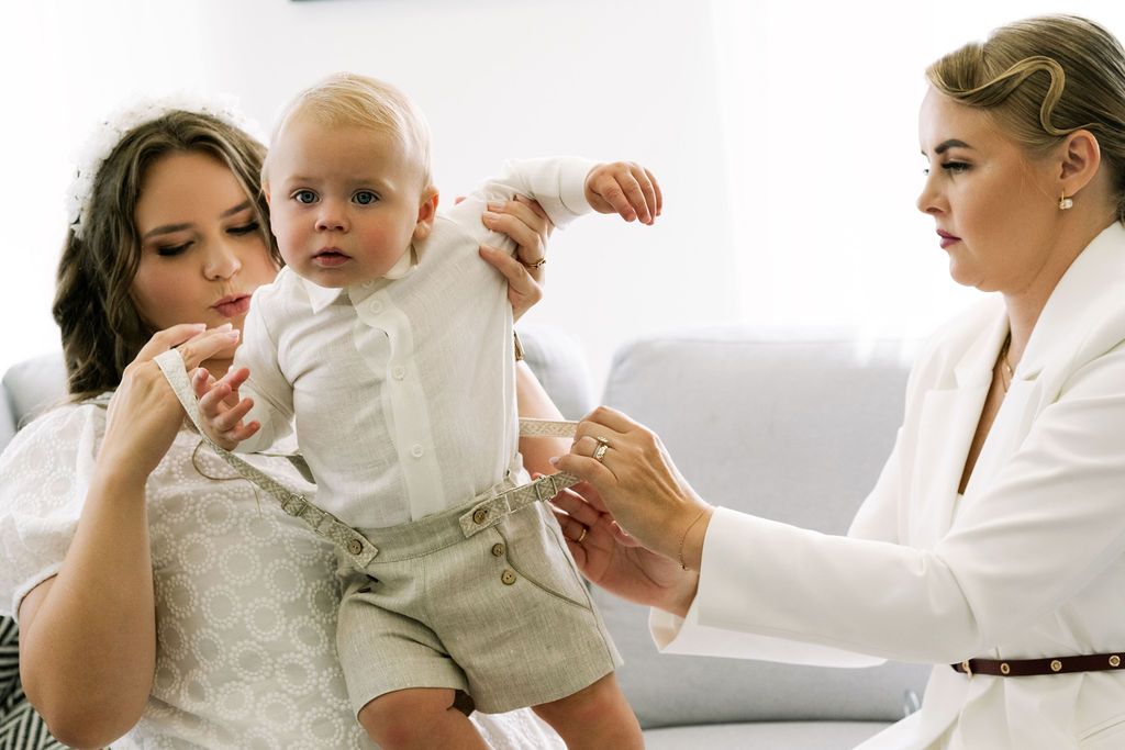 A mother holds her baby boy in white outfit while an older woman in white jacket assists during a christening ceremony.
