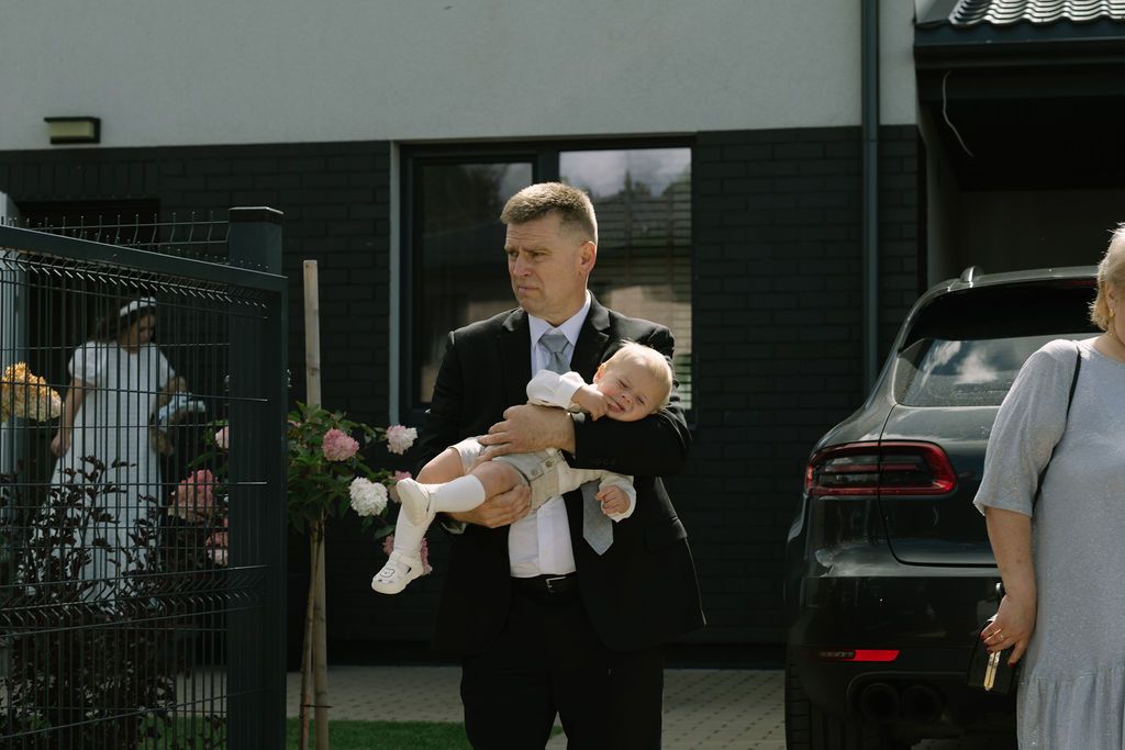 Man in black suit carrying a baby outside a modern building.