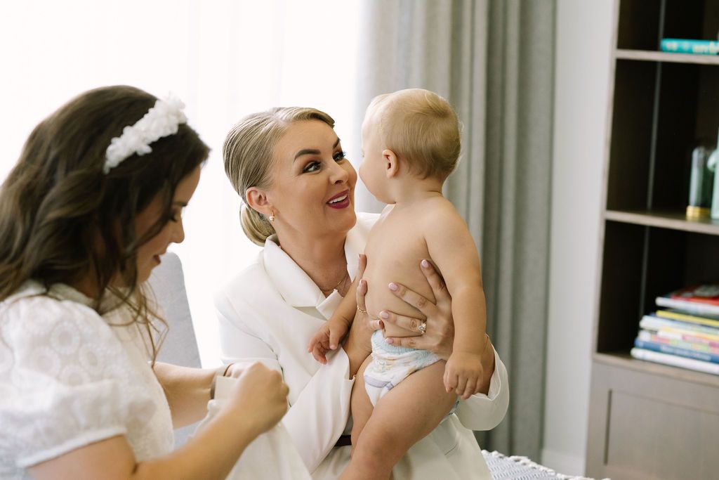 Two women in white dresses smile at an adorable baby in a bright, modern nursery room.