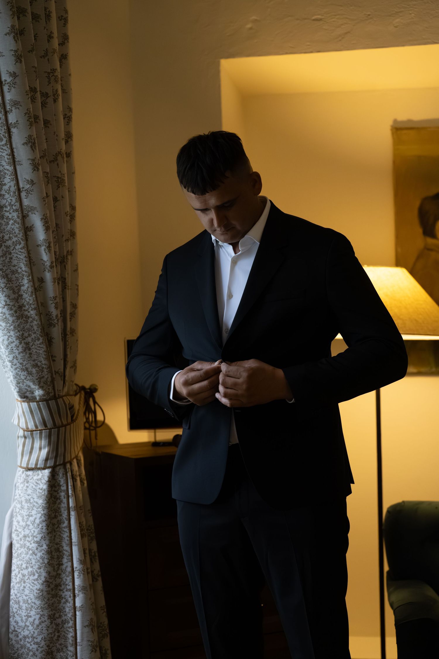 A well-dressed man in a dark suit buttoning his jacket in a warmly lit hotel room.