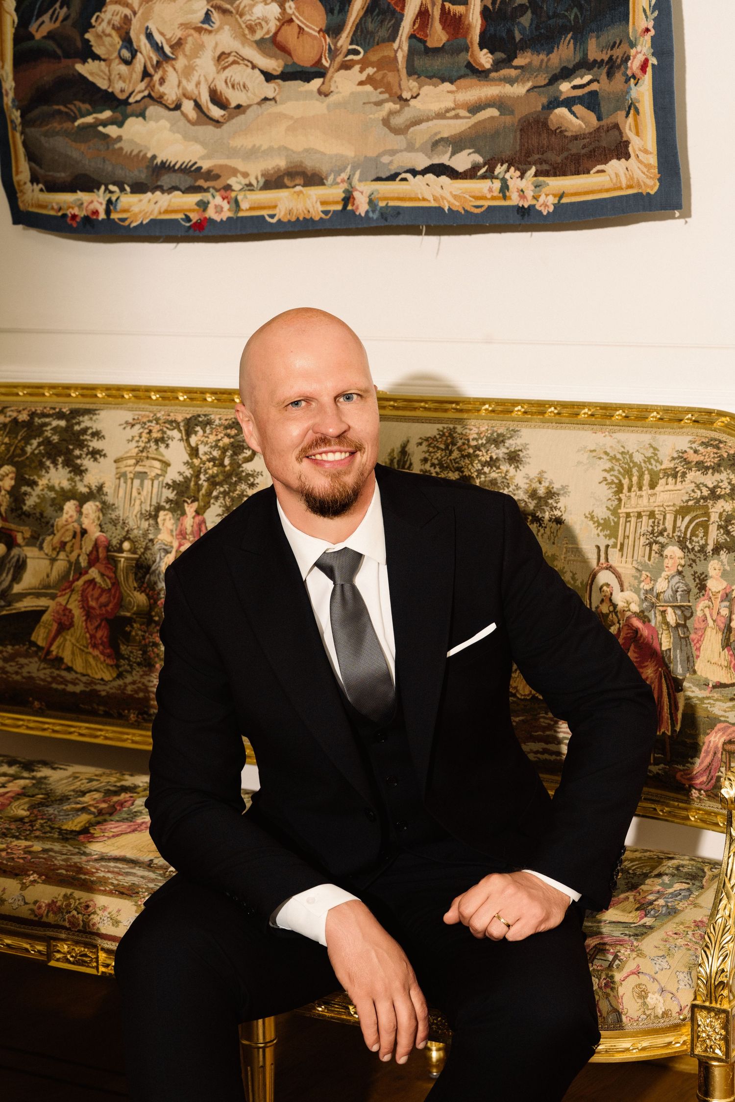 Smiling bald man with beard in black suit and tie seated on gilded sofa with ornate tapestry backdrop.