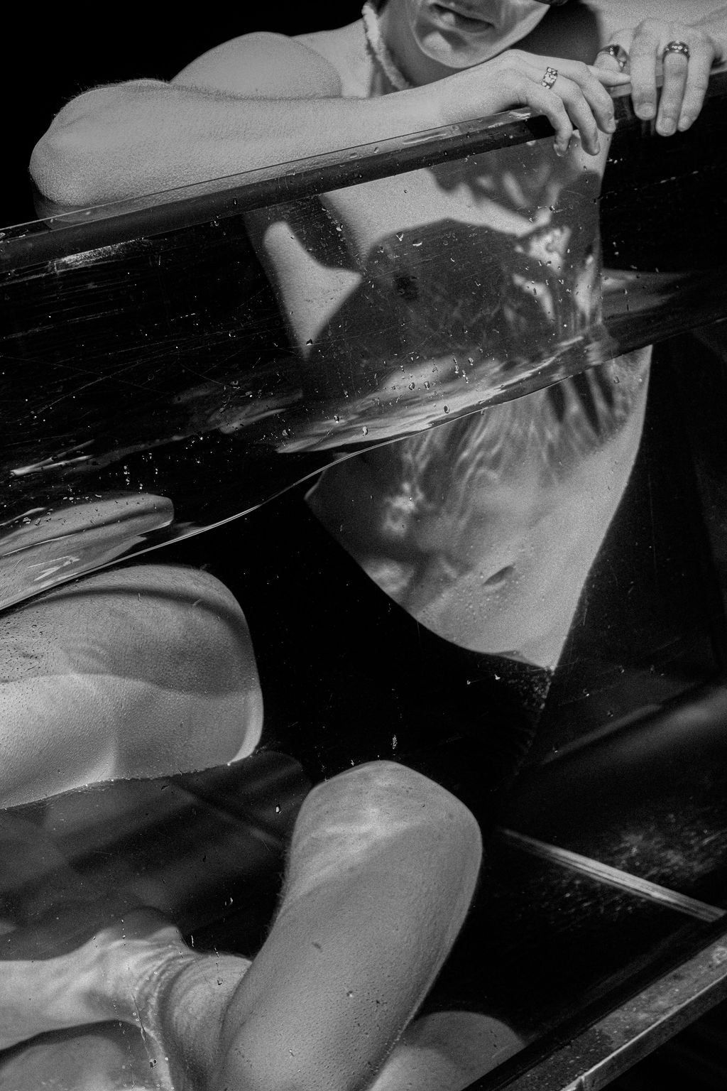 Black and white photo of a person in a dark swimsuit leaning on a glass tank rail, with their legs visible underwater below the surface.