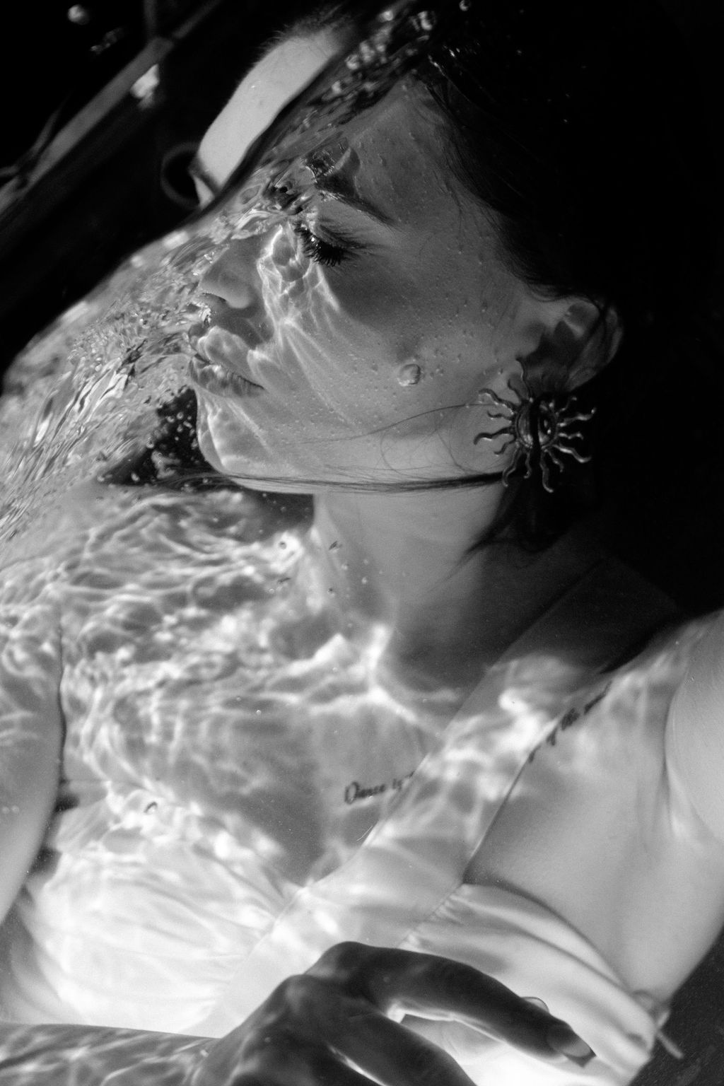 Black and white photo of a woman partially submerged in water, sunlight rippling across her face, with a sun-shaped earring.