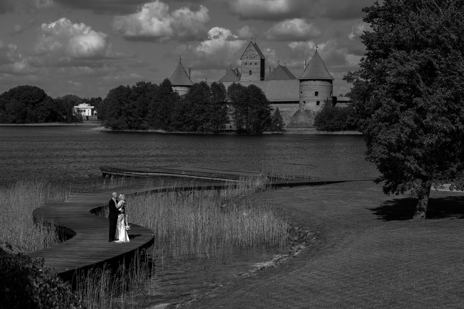 Black and white photo of a bride and groom standing on a curved lakeside path with a historic island castle in the background.