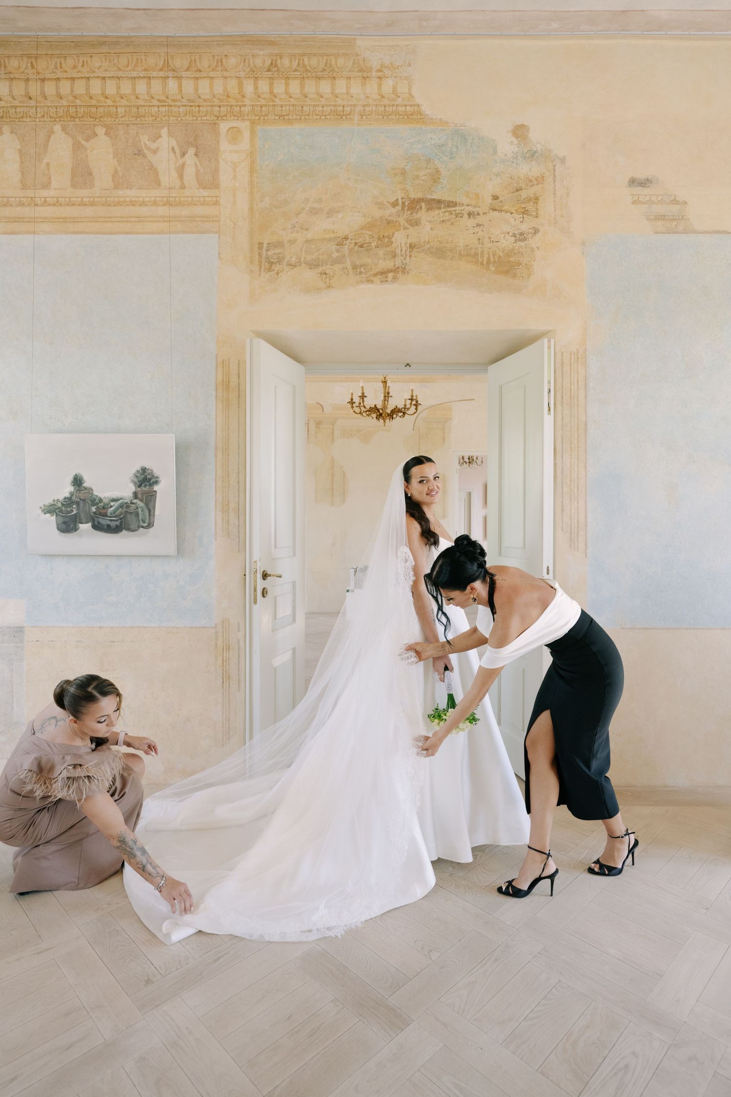 A bride in a long-veiled wedding gown is helped by two women in a frescoed room with tall white doors and a chandelier.