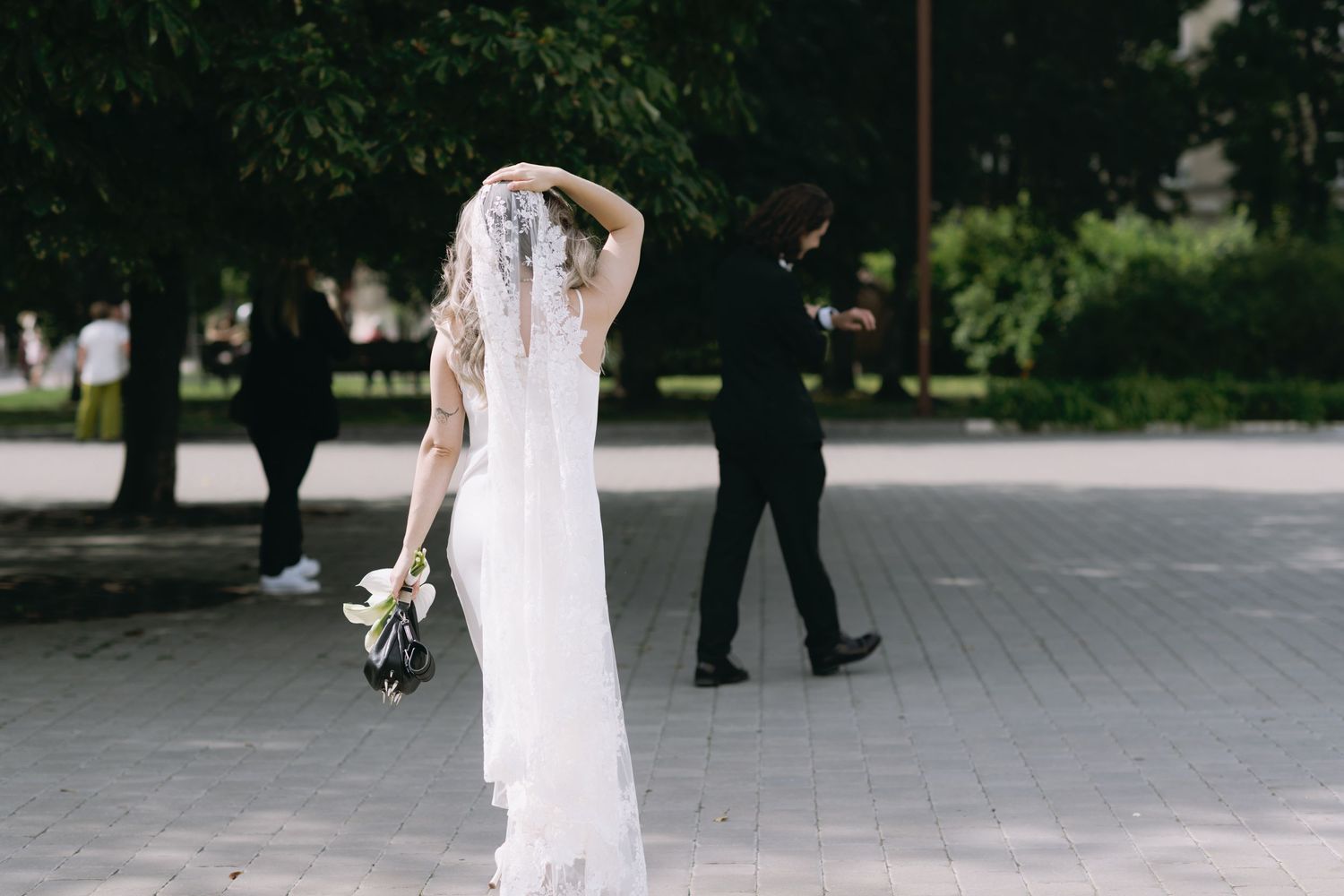 Bride in a long white dress and lace veil holding a bouquet while walking on a sunlit paved path toward a man in a dark suit.