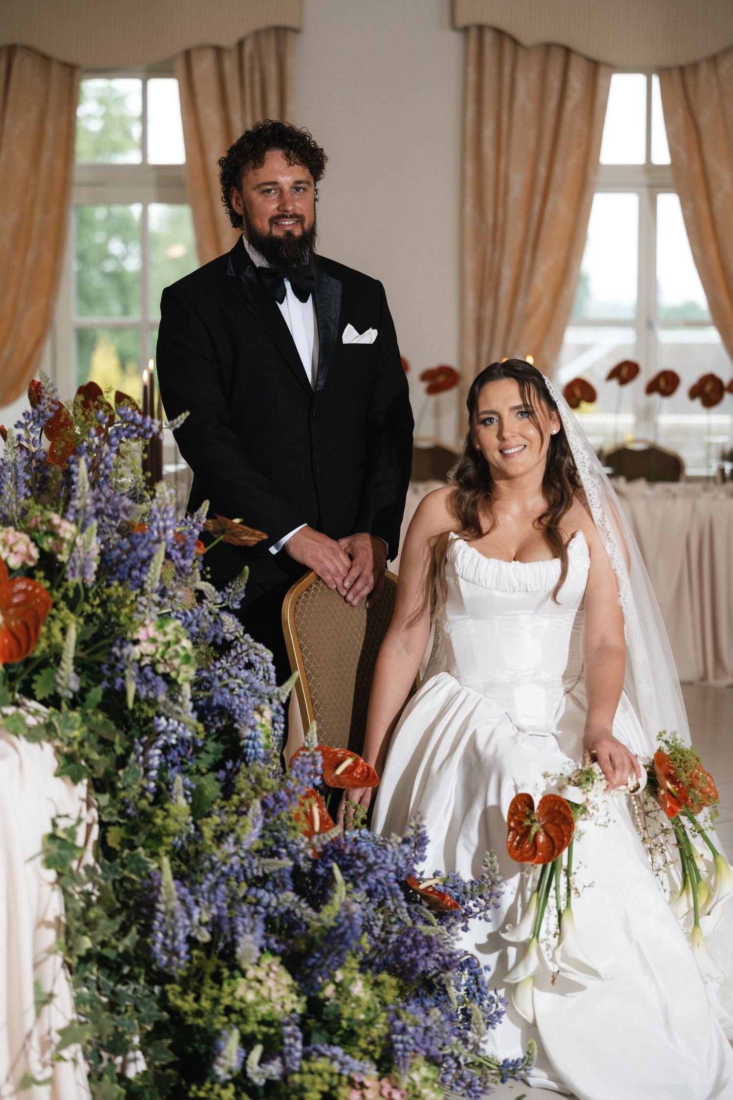 Bride in white gown and groom in black tuxedo pose beside blue and red floral arrangements at elegant wedding reception.