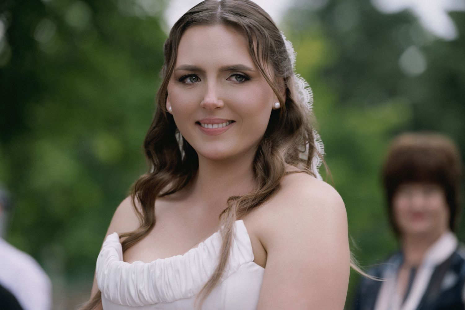 Beautiful bride in a white strapless ruffled gown smiling outdoors with soft green foliage in the background.