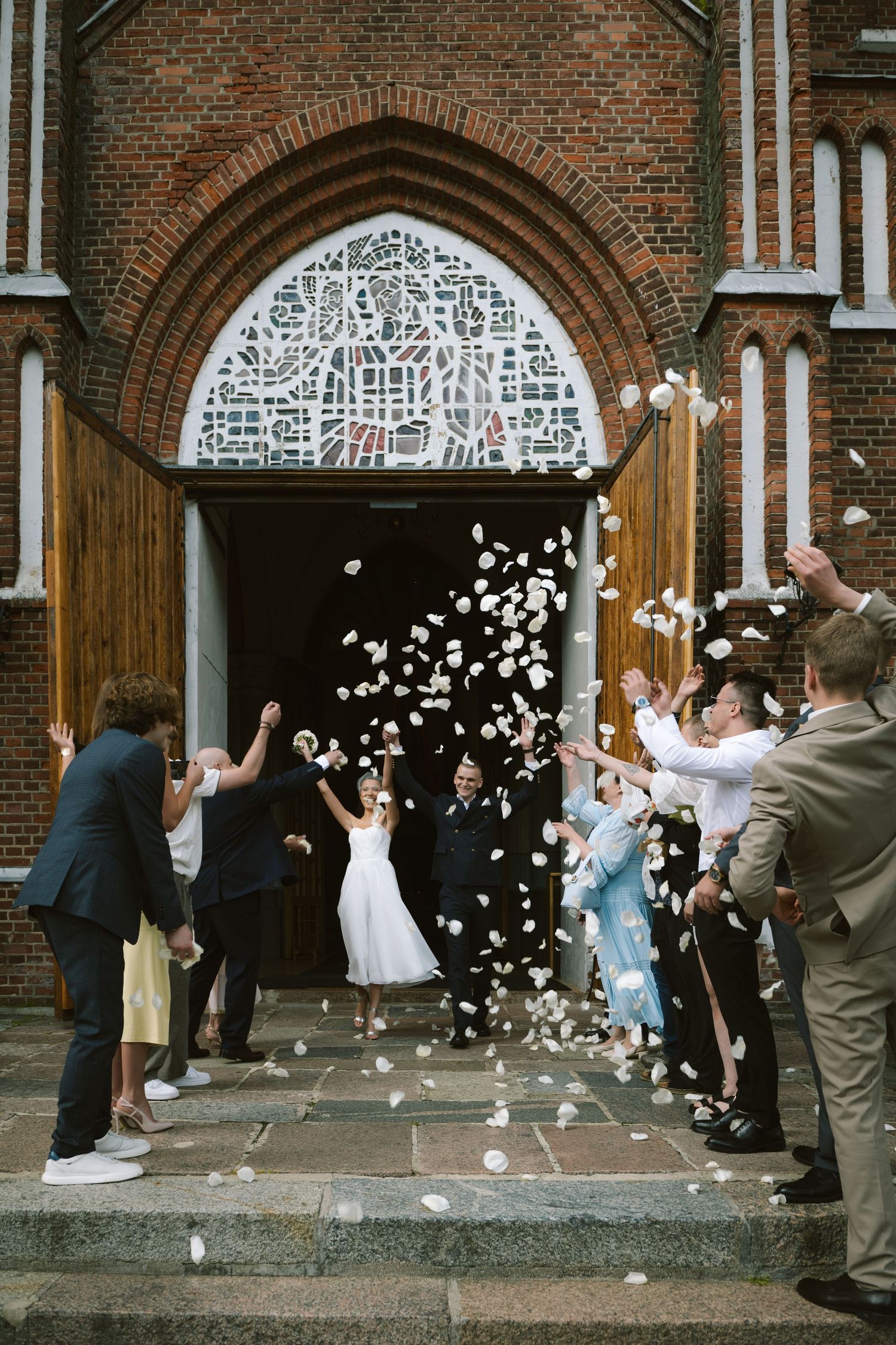 Bride and groom exit brick church as guests toss white flower petals in joyful wedding celebration.