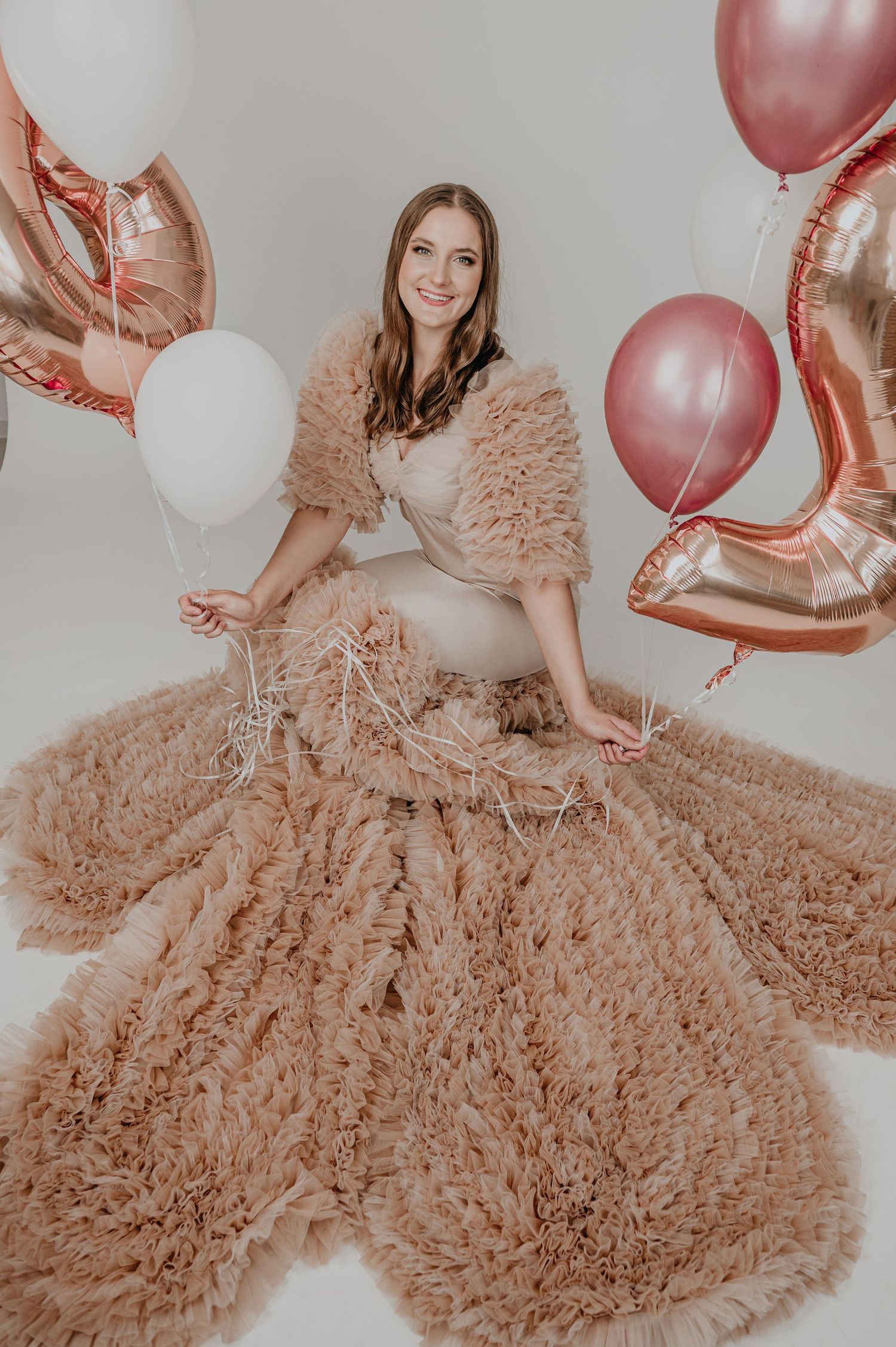 Woman in fluffy tan gown sits surrounded by rose gold and white balloons in a bright studio setting.