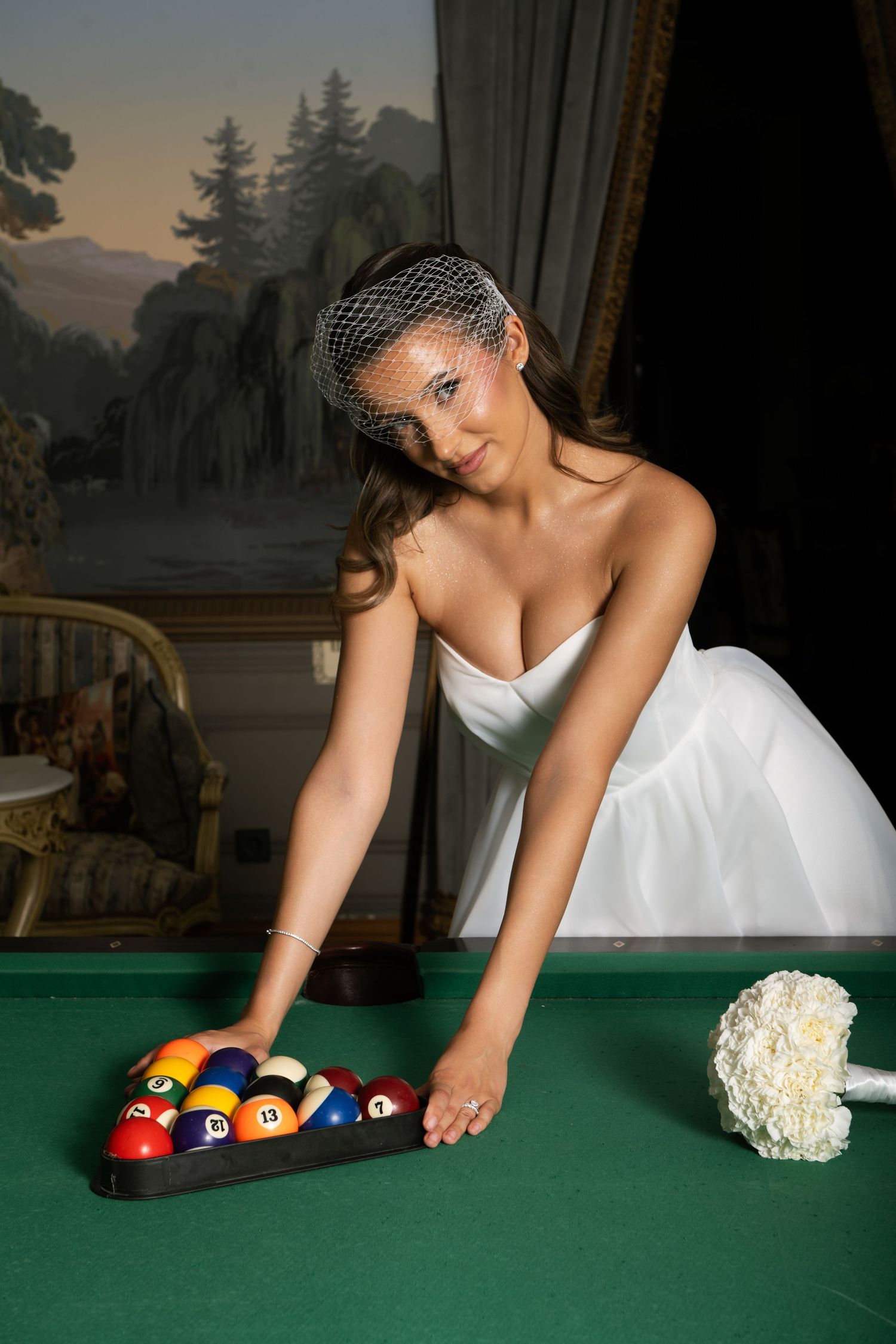 Bride in white gown and veil arranging billiard balls on a pool table with a white bouquet nearby.