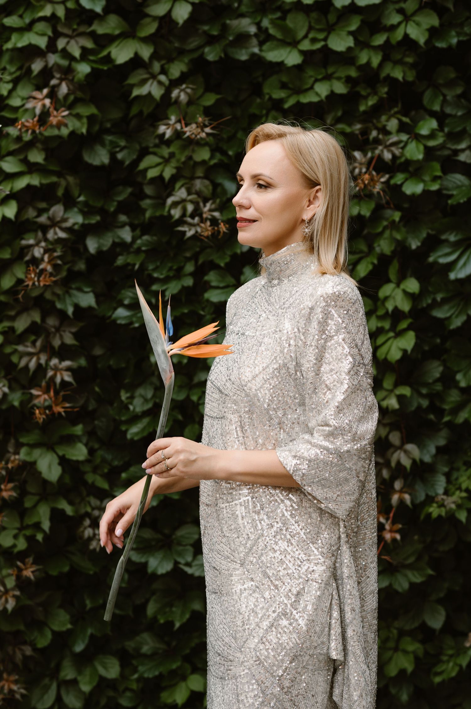 A person in a sparkly silver sequined dress poses in a garden setting against green foliage.