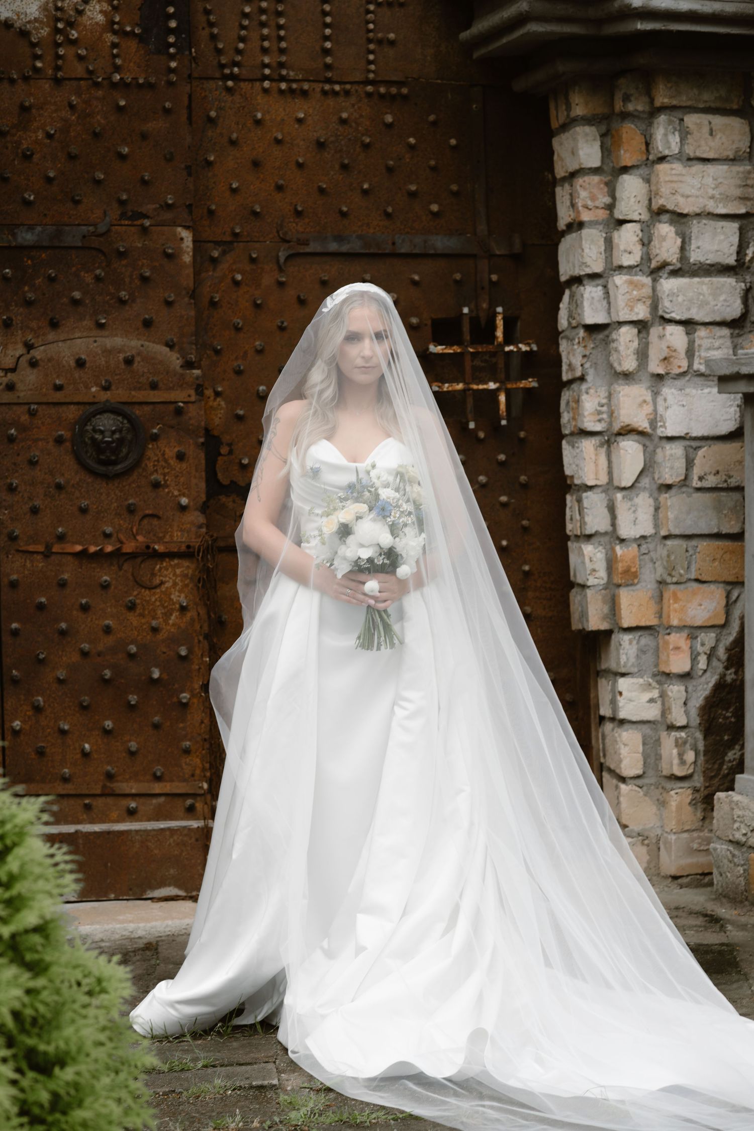 A bride in a flowing white wedding gown and veil poses against a rustic stone wall and wooden door backdrop.