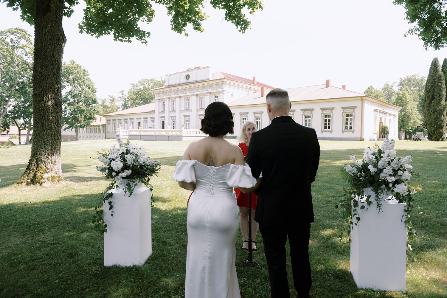 A wedding couple walks together on a grassy lawn near trees and buildings in a romantic outdoor setting.