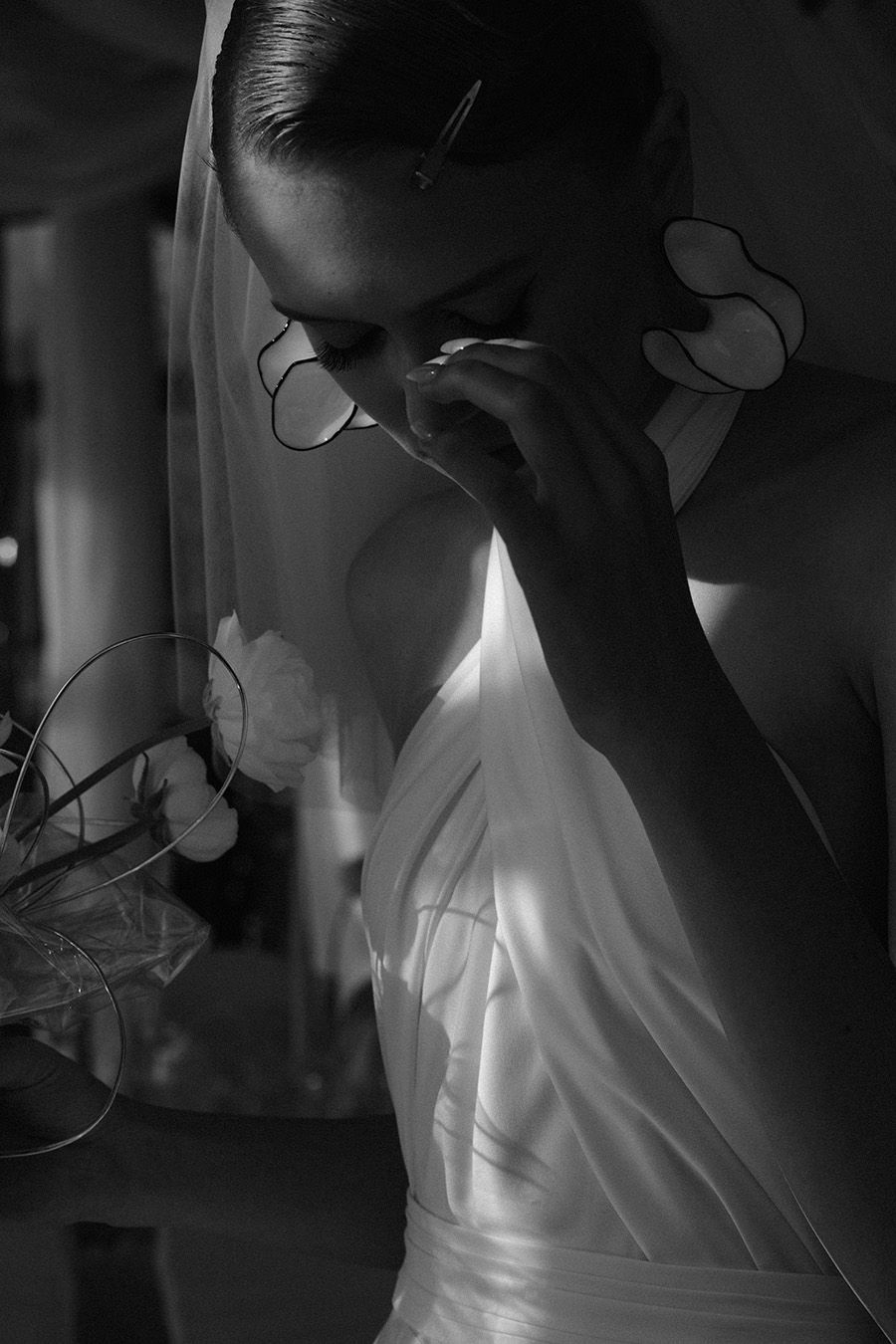 A black and white artistic photograph shows a figure in white clothing adjusting glasses in dramatic lighting.