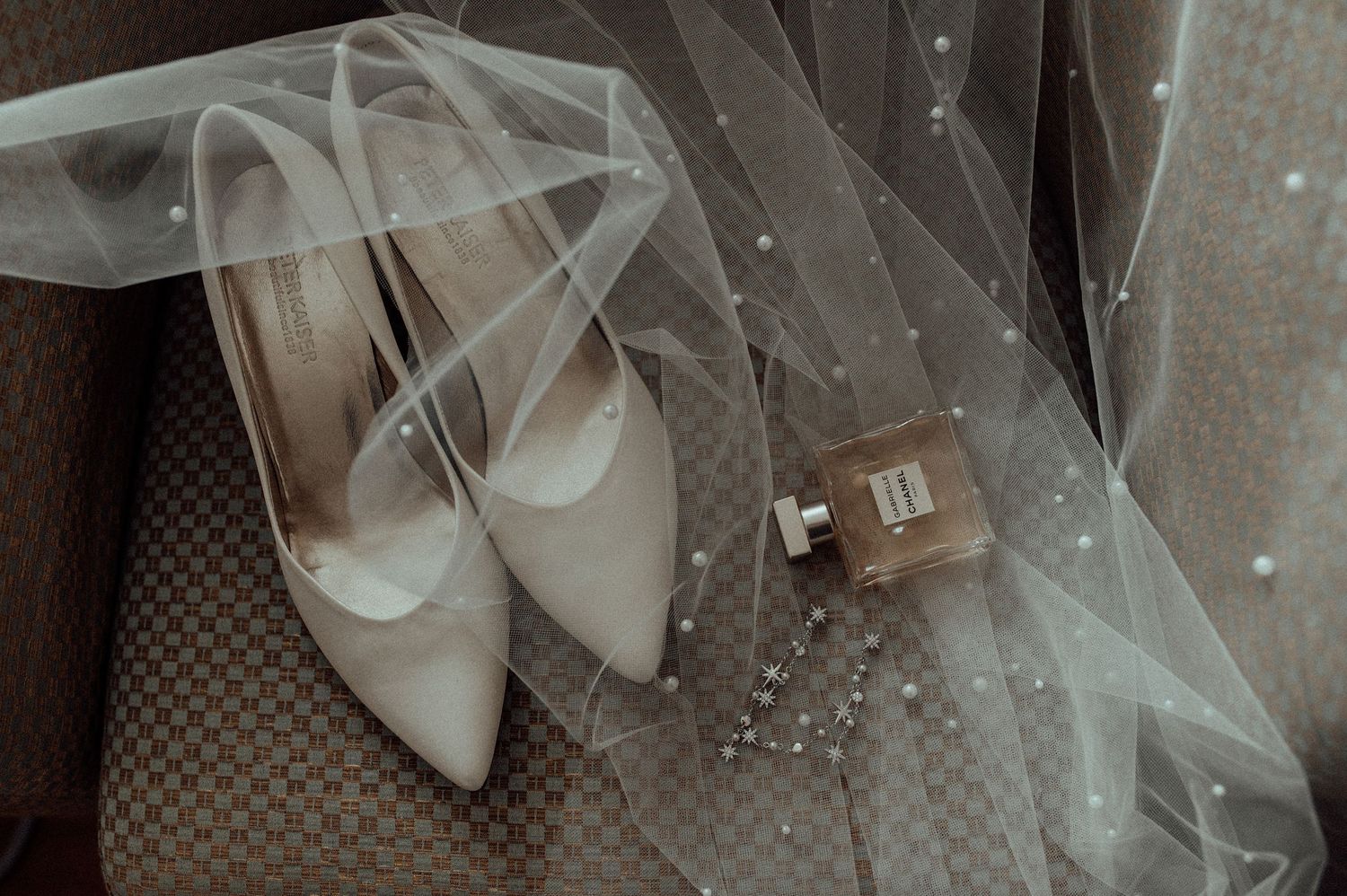 Elegant white bridal shoes and veil with scattered pearls arranged on textured gray fabric in soft lighting.