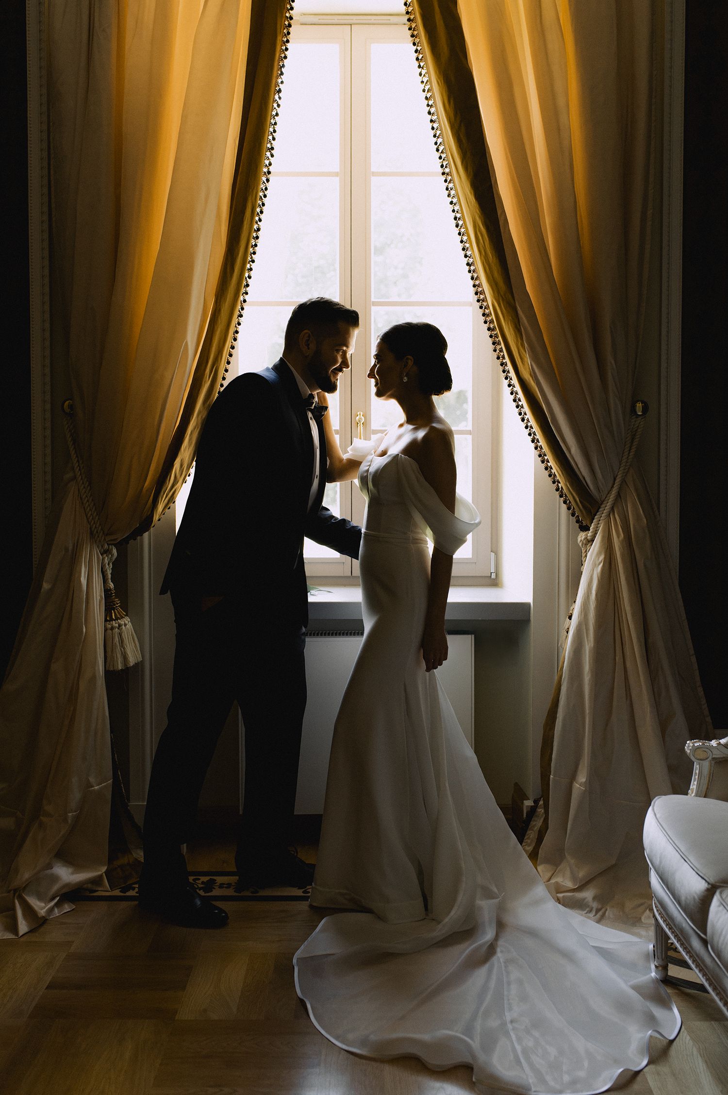 A silhouetted couple stands in front of a sunlit window with elegant gold curtains in a romantic wedding moment.