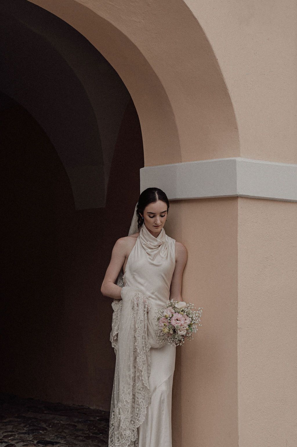 Bride in satin halter gown with lace veil holds pink bouquet under arched doorway of peach stucco building.