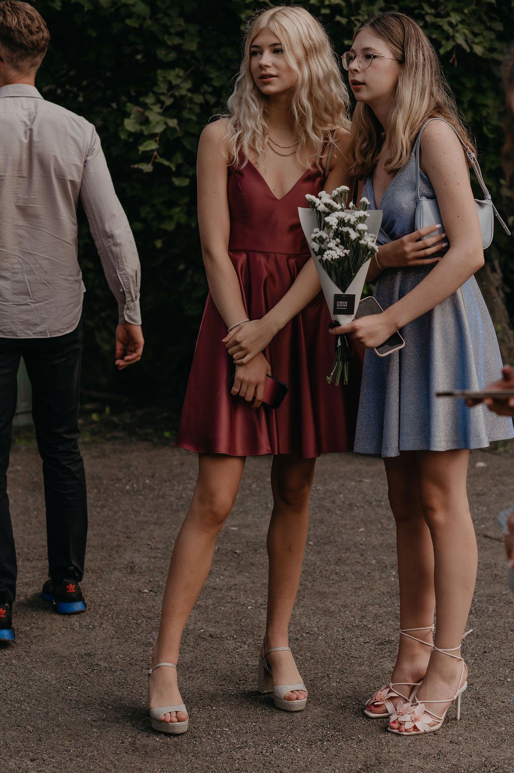 Two young women in elegant short dresses holding flowers at an outdoor event.