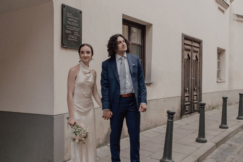 Newlywed couple holding hands in elegant wedding attire on a classic European city street.