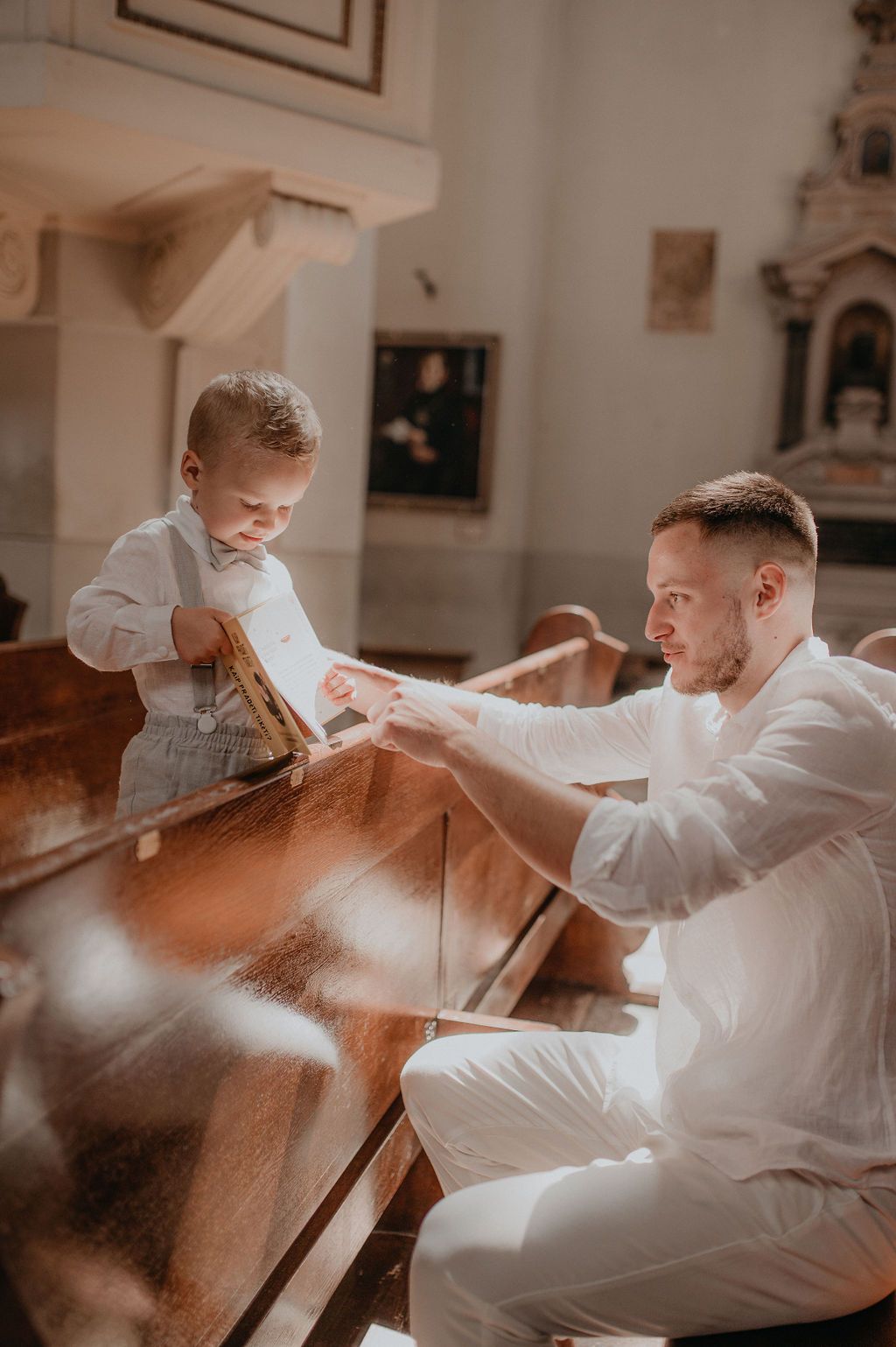 Man in white suit interacts with toddler boy in church pew during baptism ceremony.