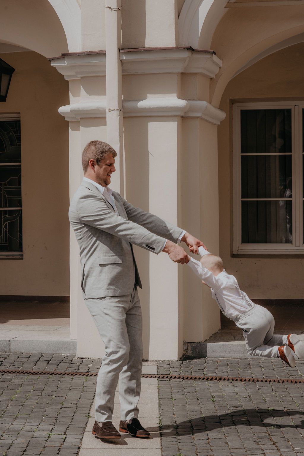 Father in light gray suit playfully swings young child by hands outside elegant building.