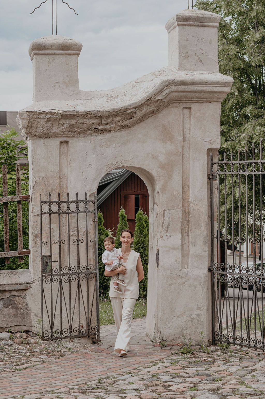 Woman carrying a baby walks through an ornate stone archway gate with wrought iron fence on a cobblestone path.