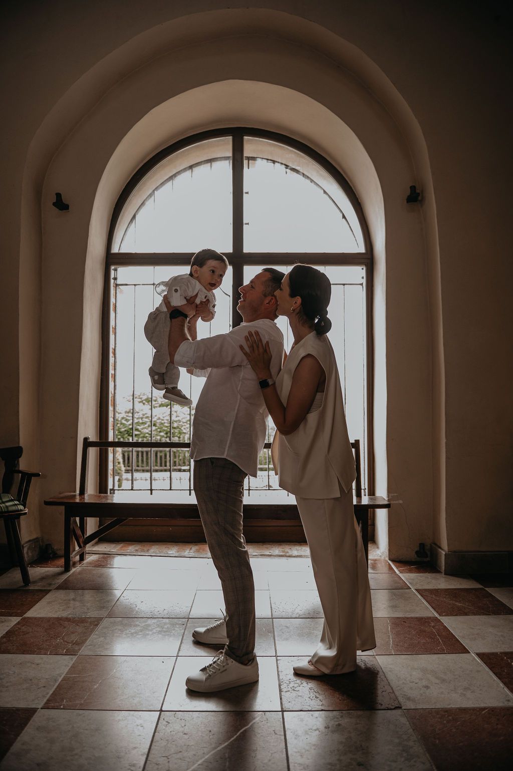 A couple in white outfits lifts their baby joyfully in an elegant arched hallway with tiled floors.