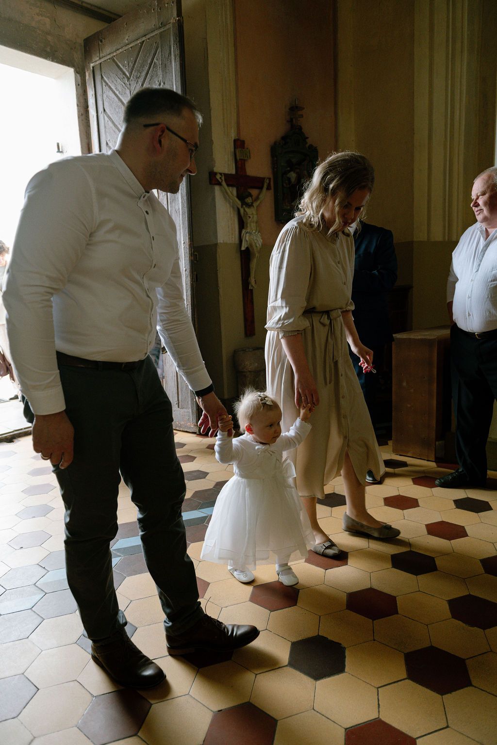 Family with toddler in white dress walking on hexagonal tile floor inside a church during a baptism ceremony.