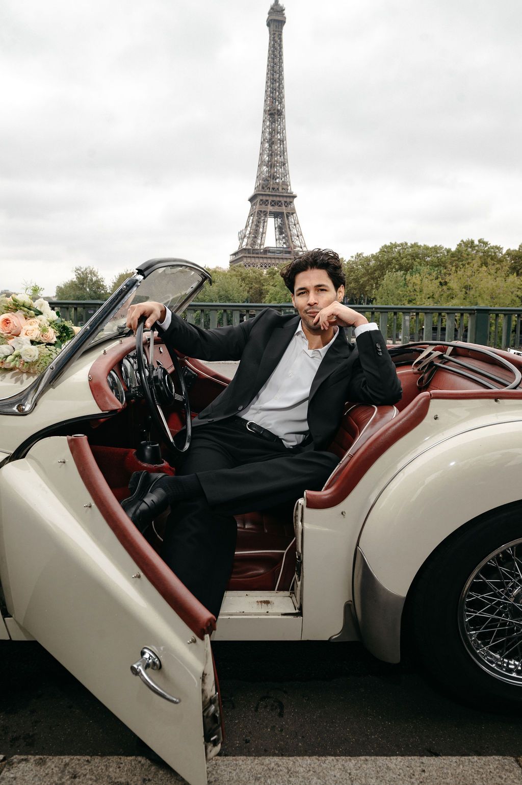 Elegant man in black suit sits in vintage convertible car with Eiffel Tower in Paris behind him.