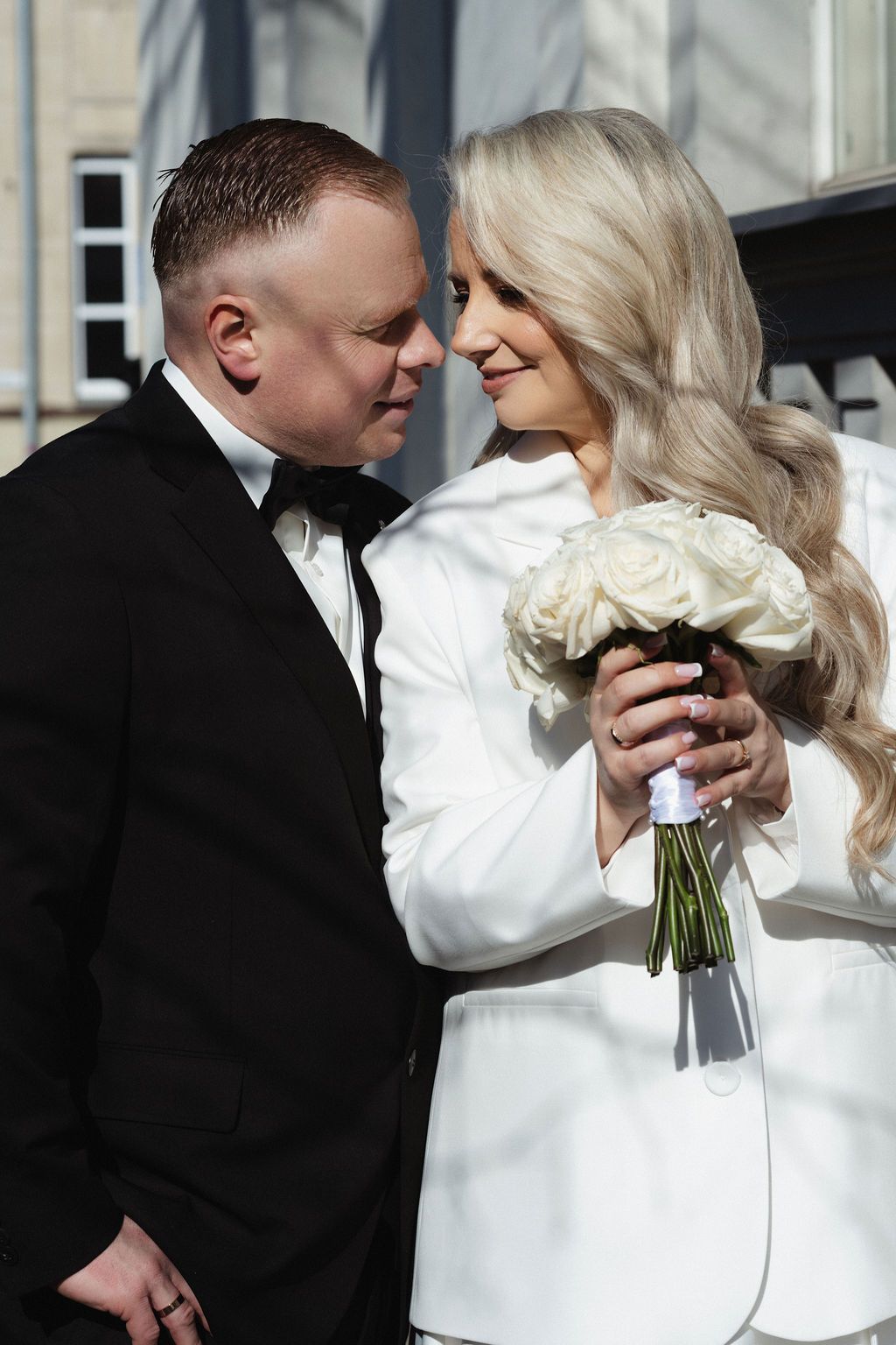 A bride in white holding roses and groom in black tuxedo share a romantic moment outdoors on their wedding day.
