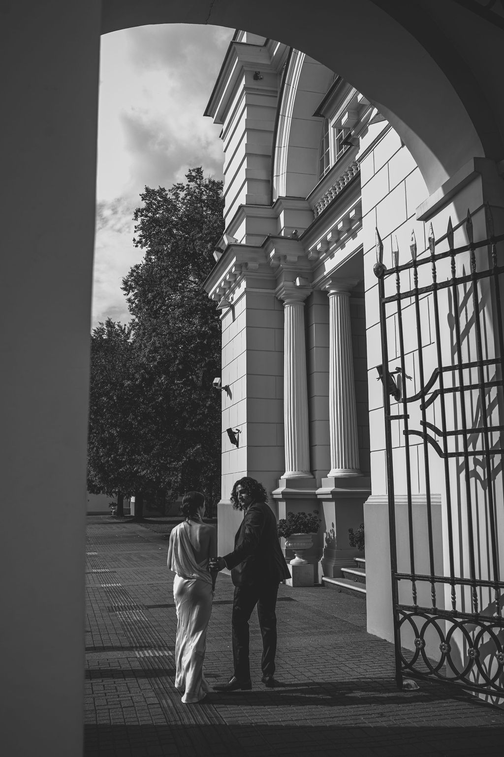 Black and white photo of a couple holding hands under an ornate archway beside a classical columned building.