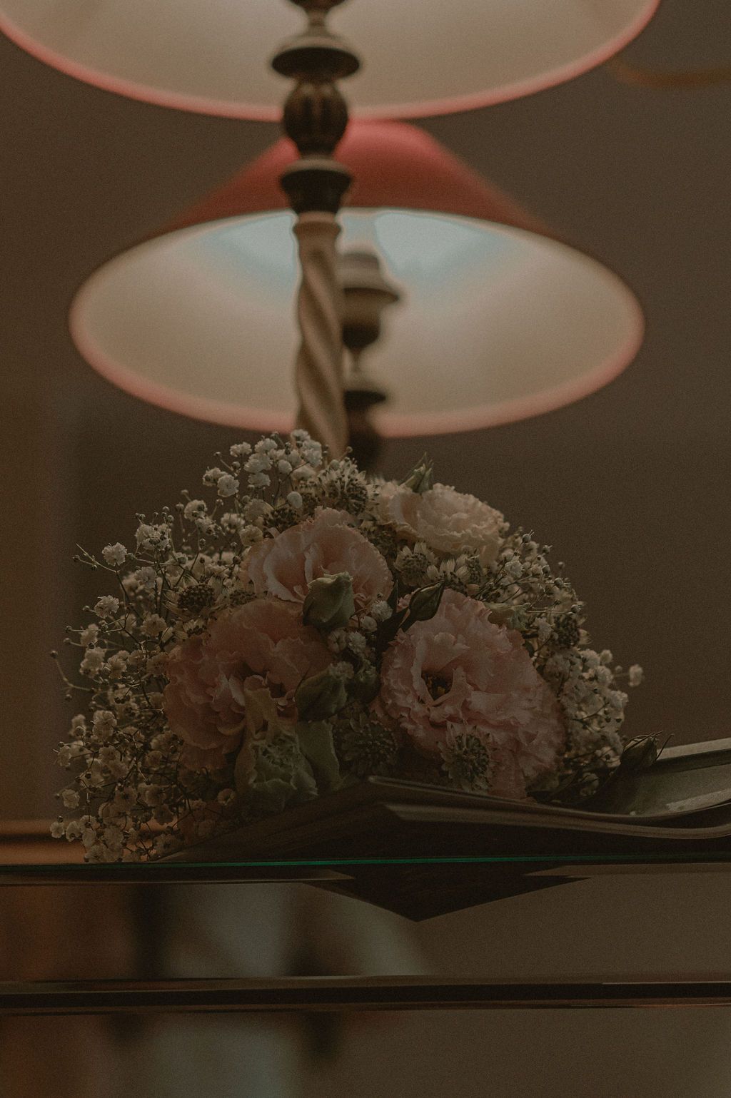 Elegant floral bouquet of pink roses and white baby's breath on a glass surface beneath warm glowing pendant lamps.