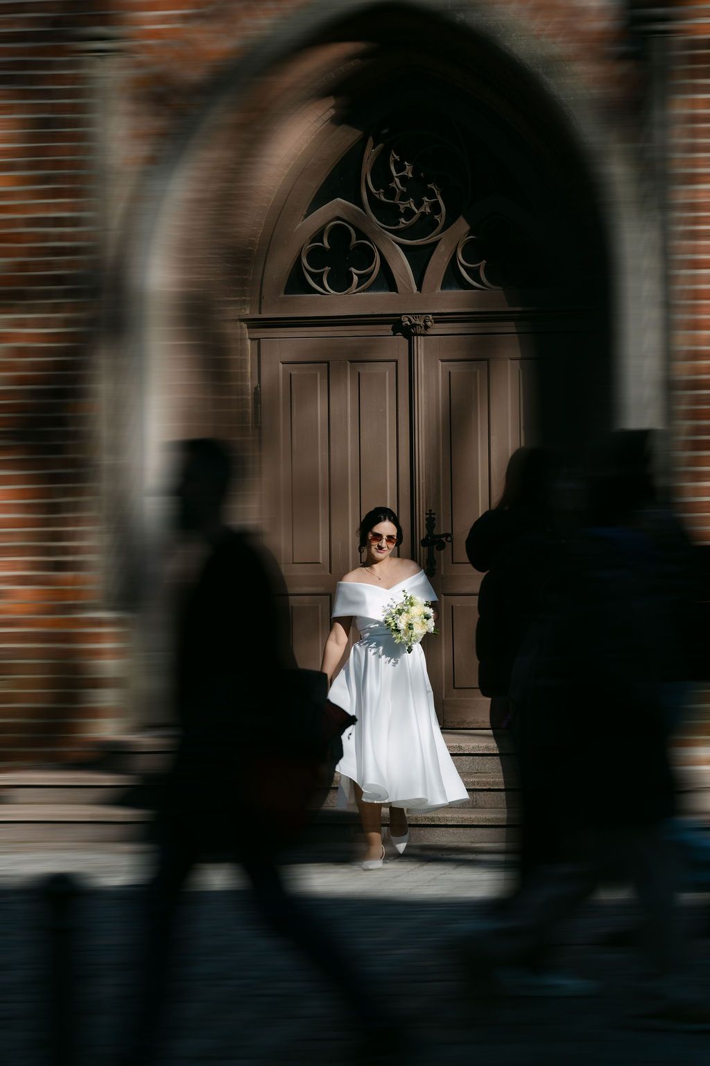 Bride in white dress holding bouquet stands before ornate church door, framed by blurred silhouettes of passersby.