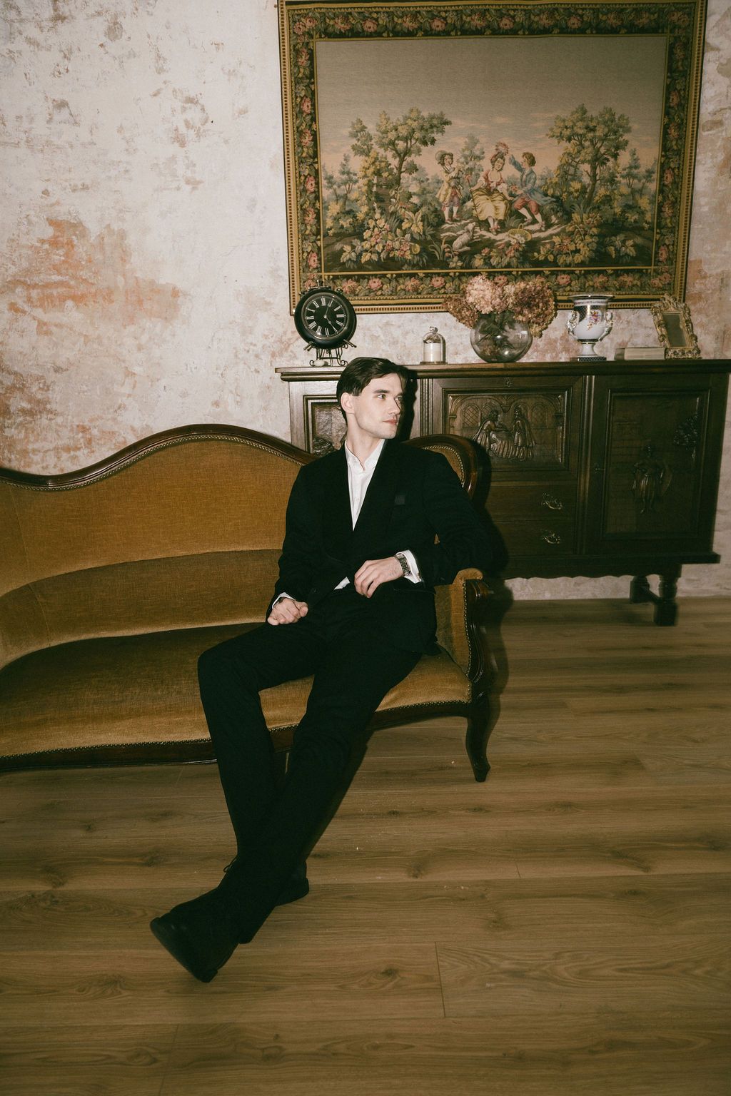 A young man in a black suit sits on a vintage sofa in an elegant room with antique furniture and tapestry.