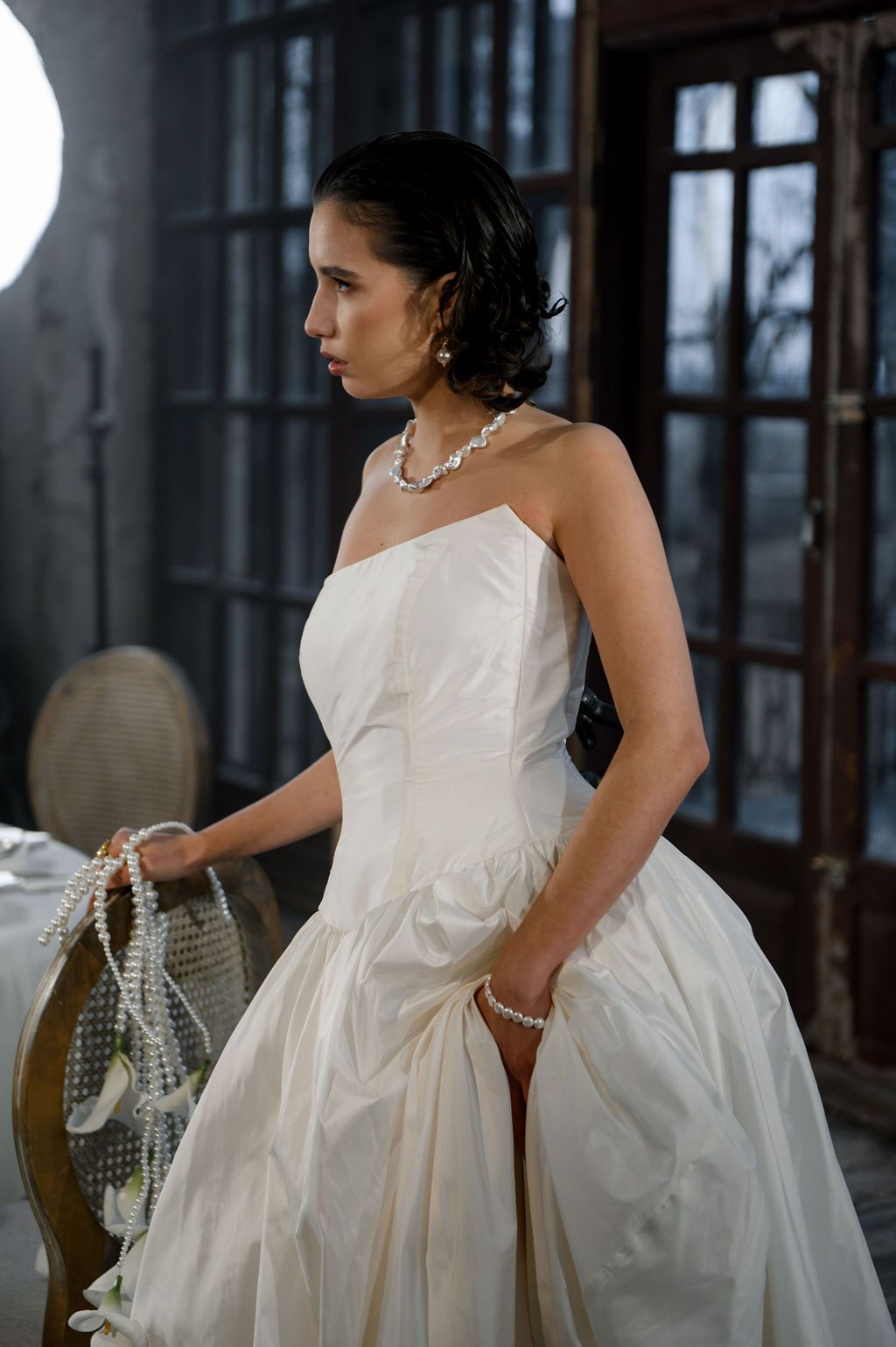 Elegant bride in strapless white ball gown wearing pearl jewelry, posing indoors in moody dramatic lighting.