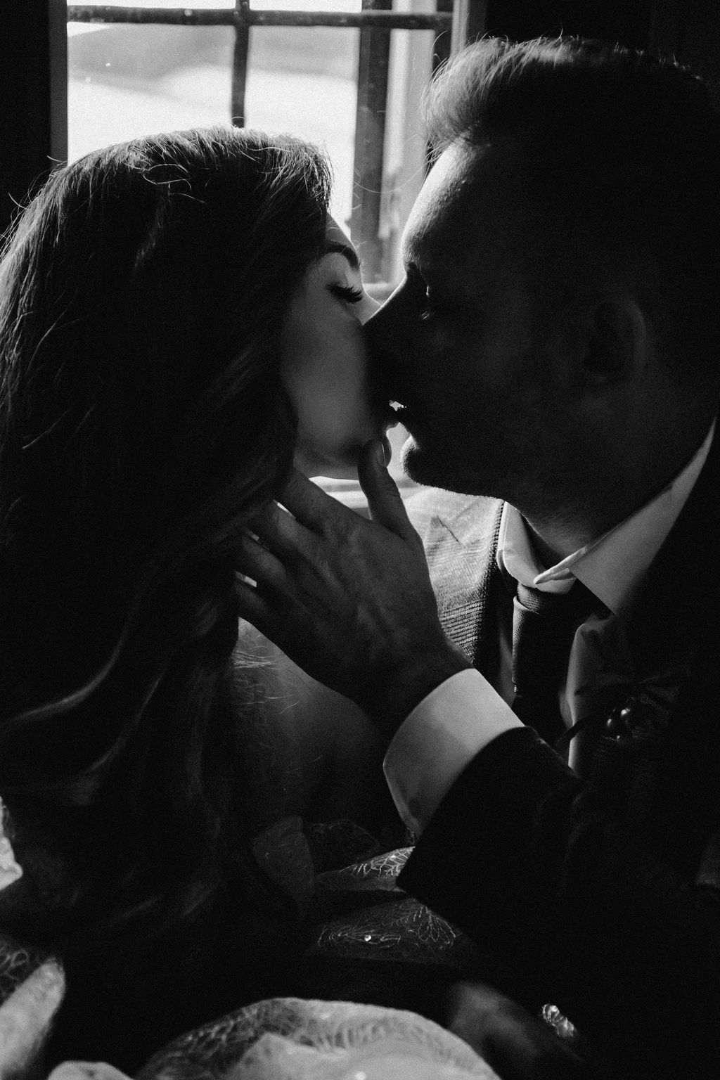 Black and white photo of a bride and groom sharing a romantic kiss near a window on their wedding day.
