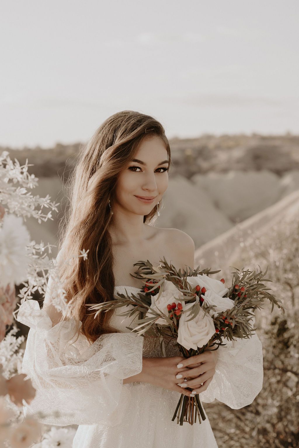 Bride in off-shoulder lace gown holds bouquet of white roses and red berries outdoors in soft natural light.