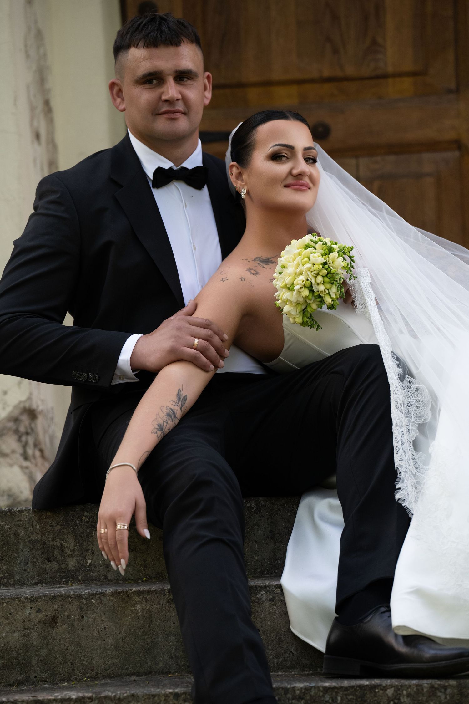 A bride and groom sit on stone steps, she holds white flowers, wearing a veil, he wears a black tuxedo with bow tie.
