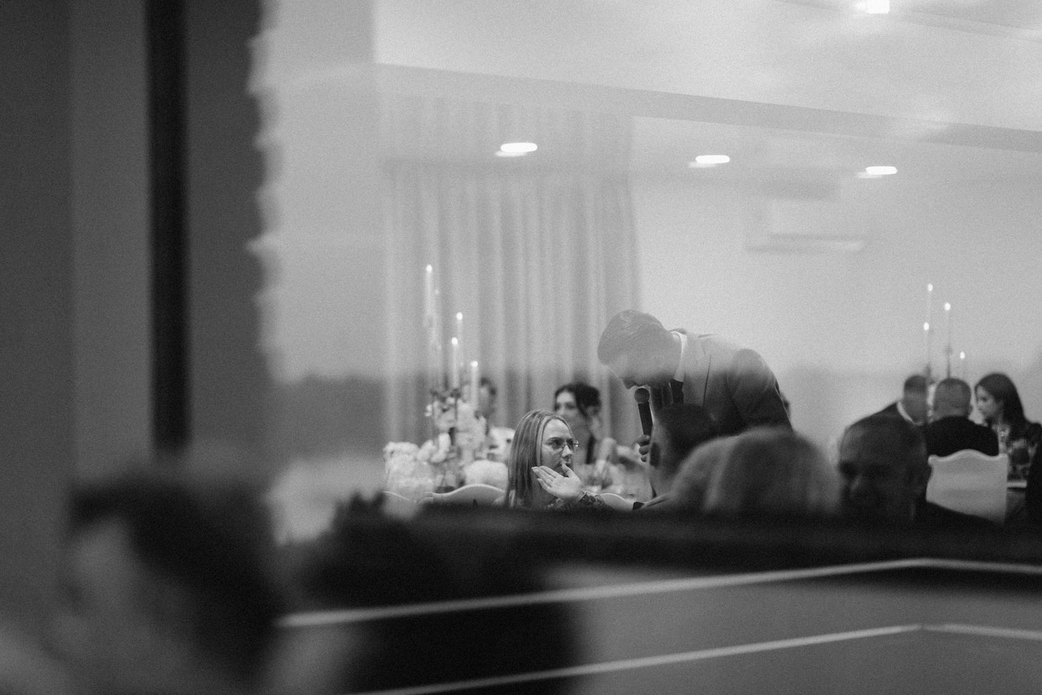 Black and white photo of guests seated and chatting at a banquet reception, viewed through a window with soft reflections.