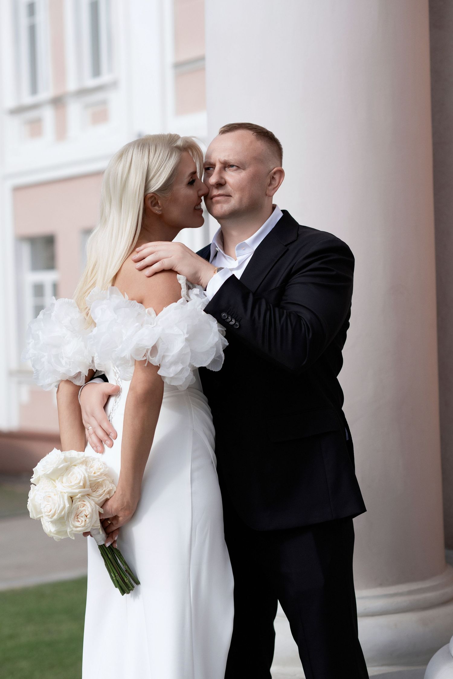 Bride in a white off-shoulder gown with ruffled sleeves holding white roses embraces a groom in a dark suit beside a classic white column.