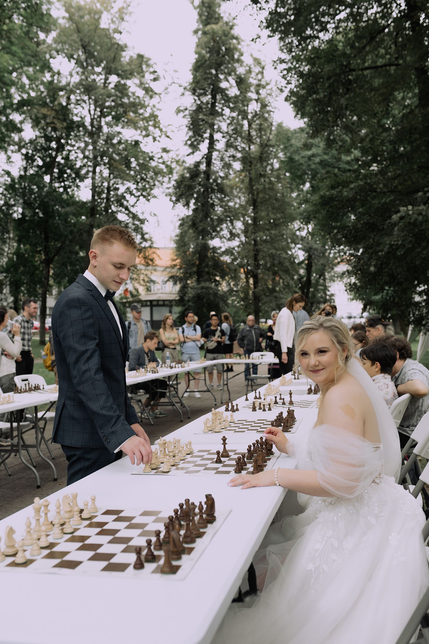 A wedding couple plays chess at outdoor reception with multiple chess boards set up on long white tables.