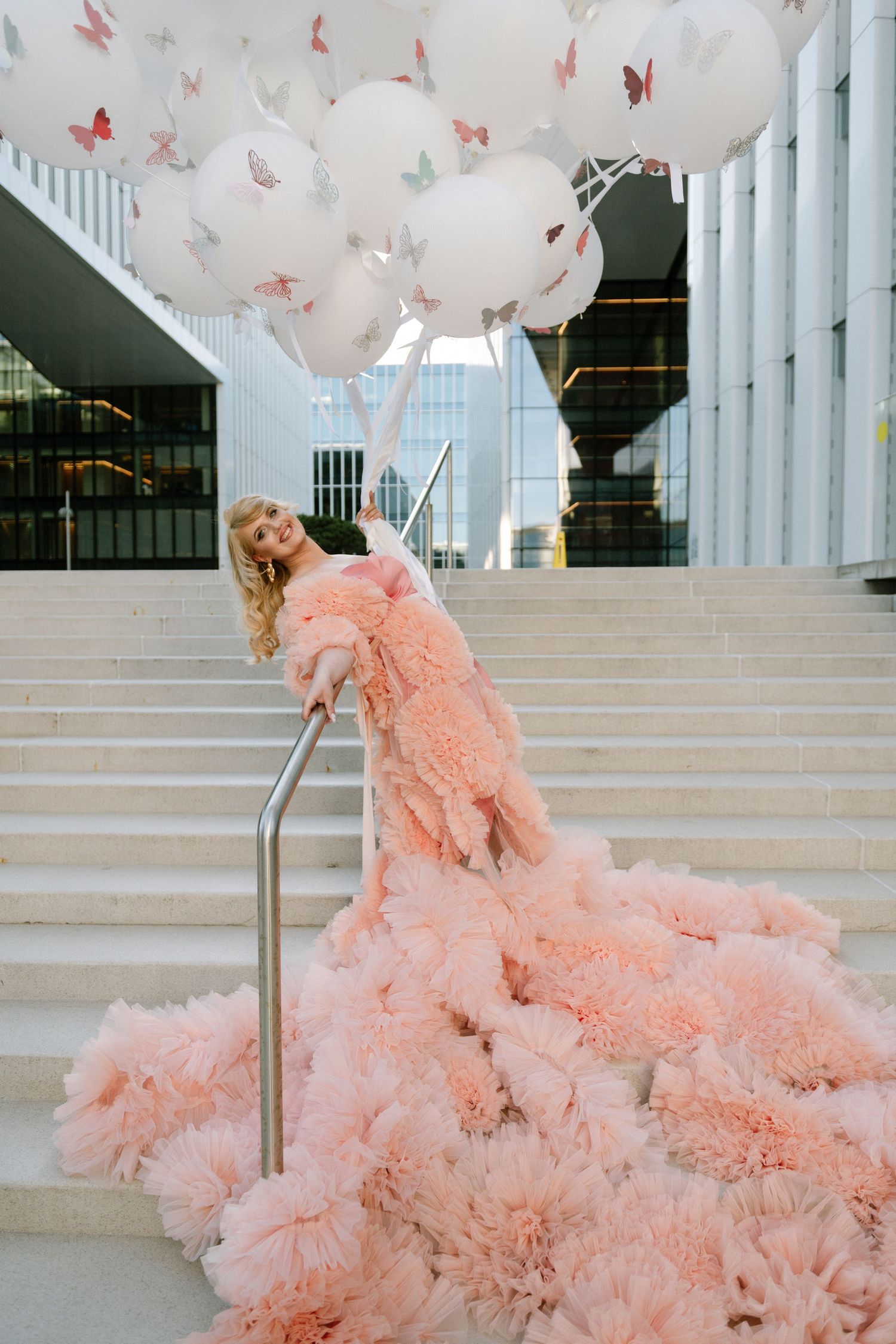 Blonde woman in a pink ruffled gown holding white butterfly balloons on outdoor stairs.