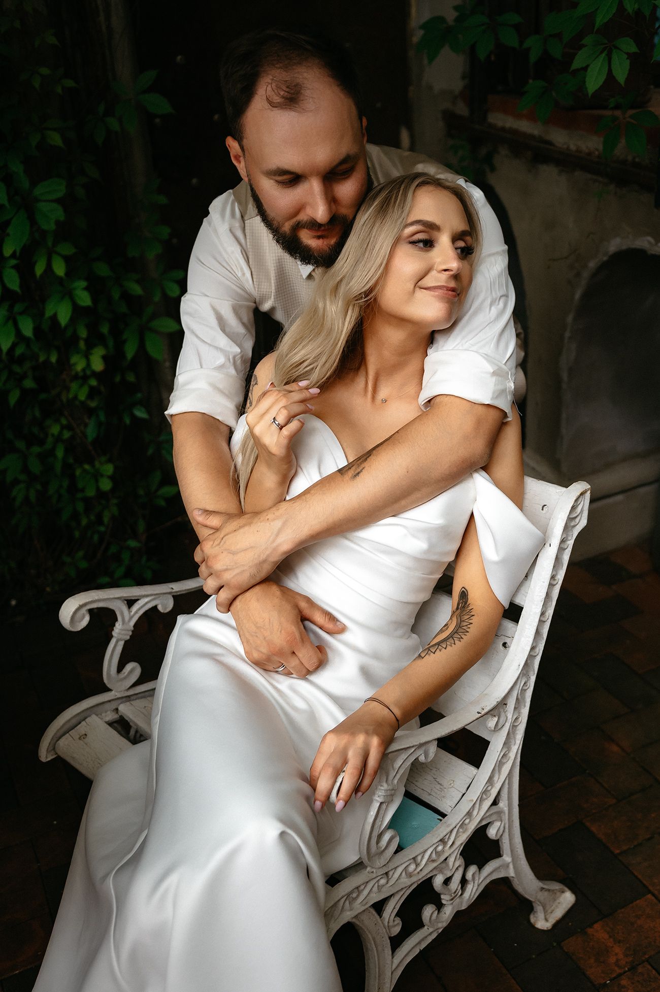 Romantic couple in white outfits share tender moments while seated on an ornate garden bench surrounded by greenery.