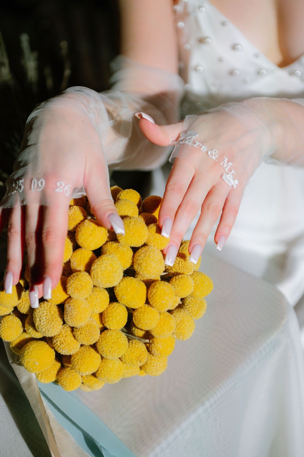 Bride in sheer lace gloves with delicate nail art holds a round bouquet of bright yellow craspedia billy ball flowers.