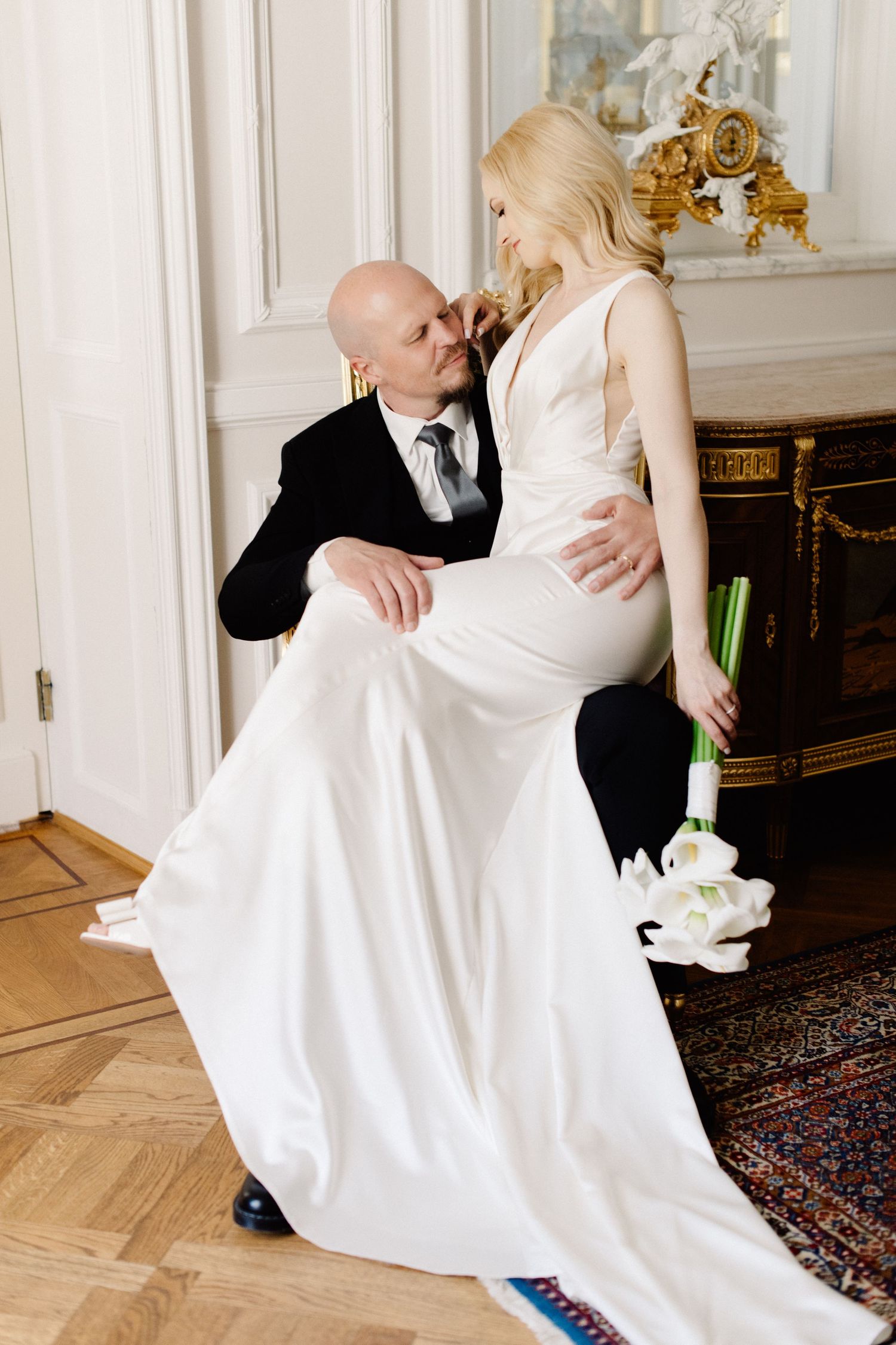 A blonde bride in a silk white gown sits on her groom's lap in a black suit, holding calla lilies in an ornate, antique-filled room.