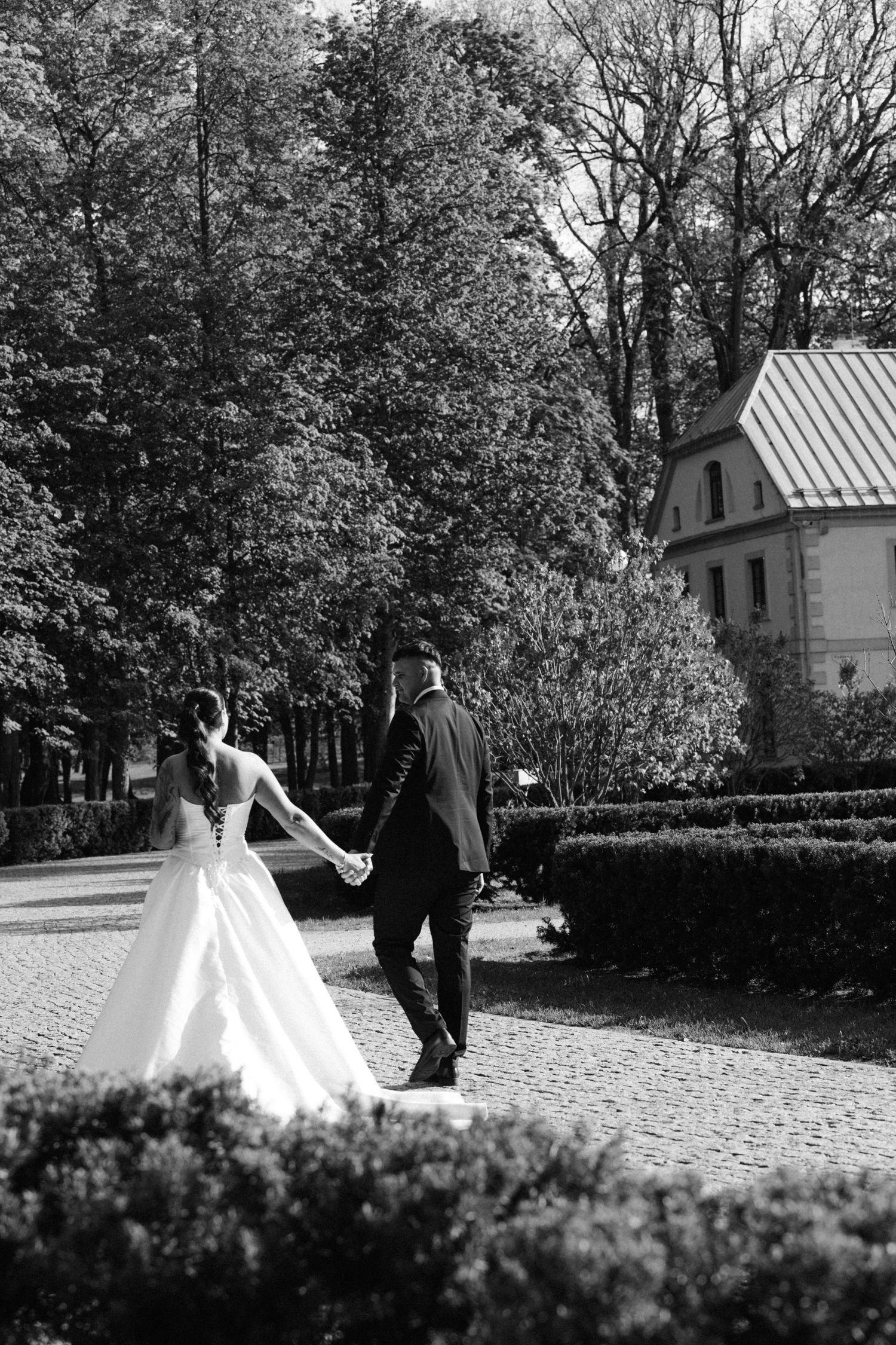Black and white photo of a bride and groom holding hands and walking along a garden path lined with hedges and tall trees.