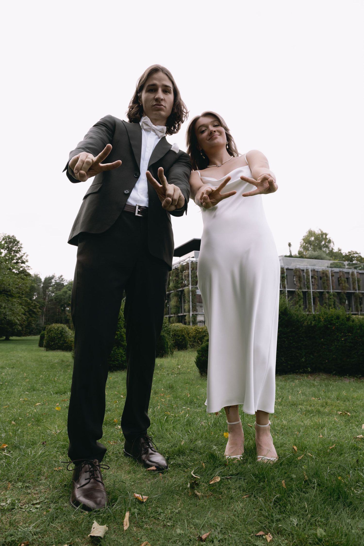 Two people in formal wedding attire make peace signs toward the camera while standing on a green lawn with hedges and trees behind them.