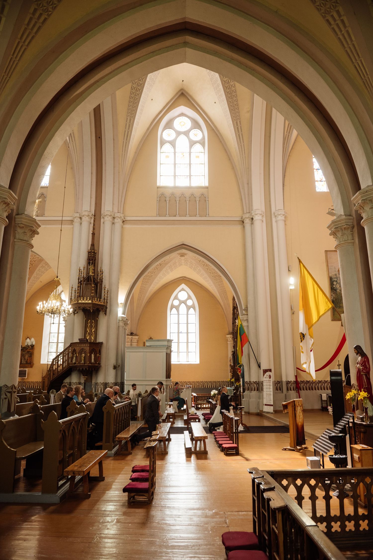 Worshippers sit in wooden pews inside a bright cream-colored Gothic church with pointed arches, tall windows and a yellow-and-white flag.