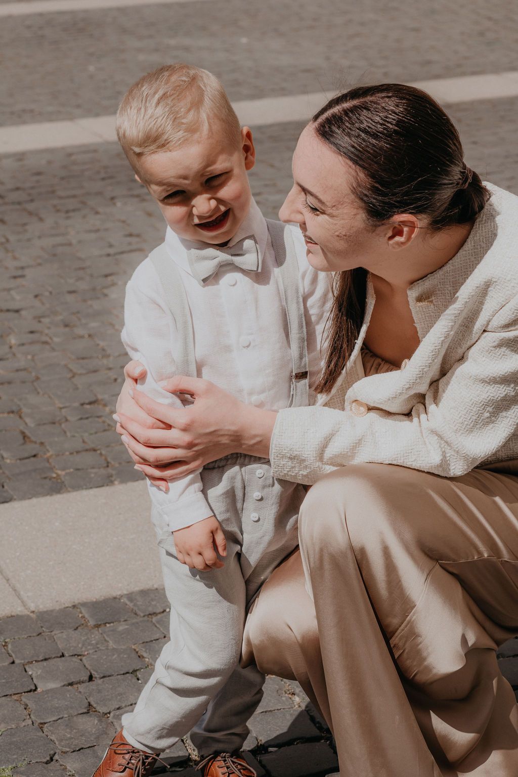 A joyful toddler in white suit laughs as a smiling woman in beige outfit crouches beside him on cobblestone.